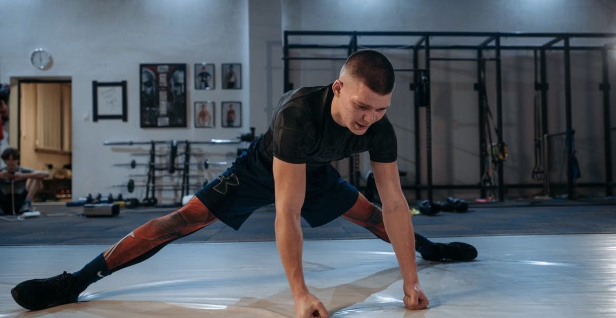 A male athlete stretches intensely in a dimly lit gym environment, preparing for training.
